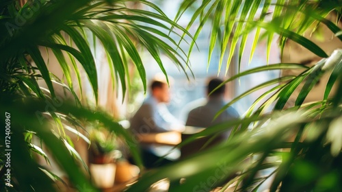 Blurred view of two people at a table, framed by lush green foliage