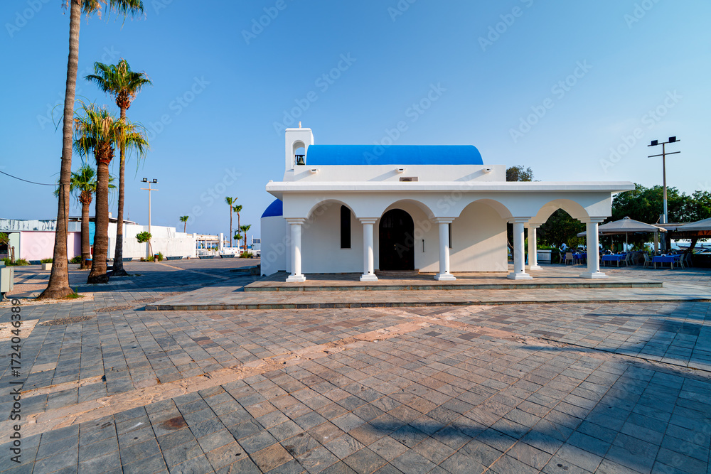 Fototapeta premium White Greek-style church building on street with palm-lined square in port Ayia Napa, Cyprus. Sightseeing in Mediterranean city on sunny day.