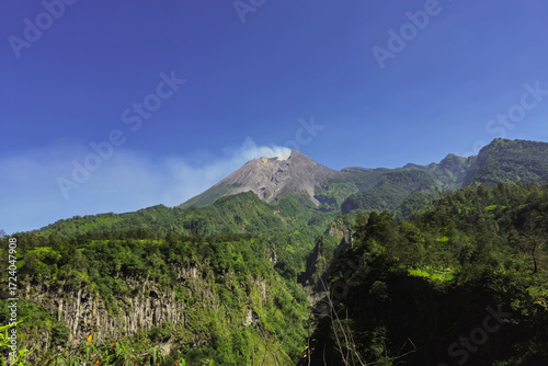 A close-up view of Mount Merapi from a distance with trees in the foreground. The panoramic beauty of Mount Merapi on a clear, midday day is clearly visible from a distance.
