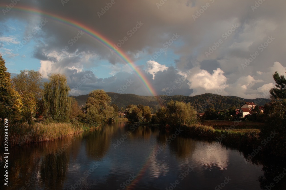 Naklejka premium Vibrant rainbow arcs across a dramatic sky over a tranquil river and lush landscape.
