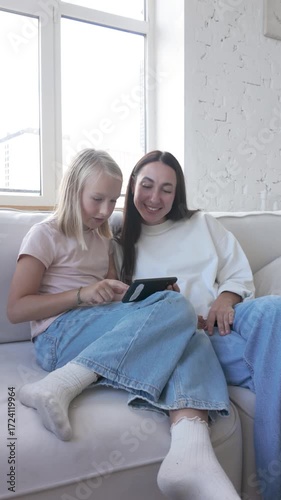 Girl shows something on smartphone to woman, they laugh and smile while sitting on the couch. Positive family moment with technology, communication and cheerful emotions at home