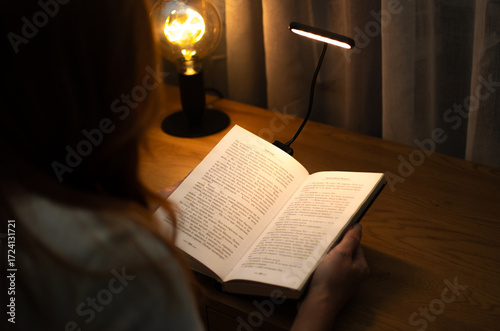 A woman is reading a book at a wooden table in the evening with the help of convenient clip-on lamp