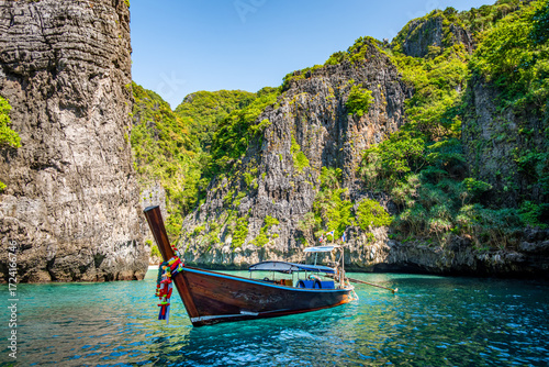 Traditional longtail boat peacefully floating on calm, clear turquoise water, surrounded by towering limestone cliffs covered with lush green vegetation on a sunny day in Ko Phi Phi Don