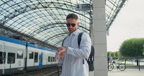 A man looks at his wristwatch standing on the station platform with a suitcase while waiting for a train.