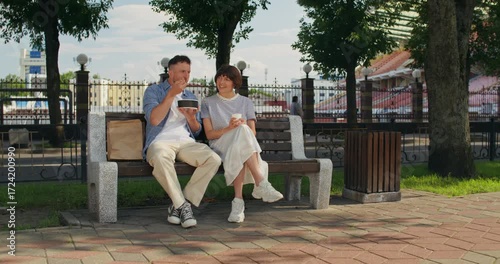 A man eats lunch sitting on a bench in the city. A woman drinks coffee sitting next to him.