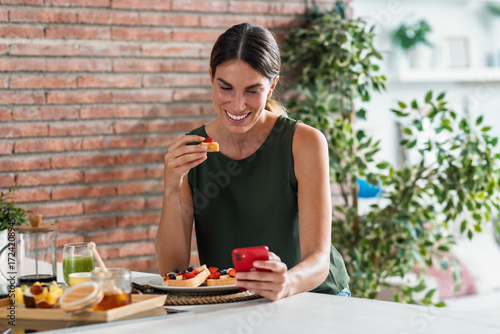 Healthy woman having delicious breakfast while using smartphone in the kitchen at home