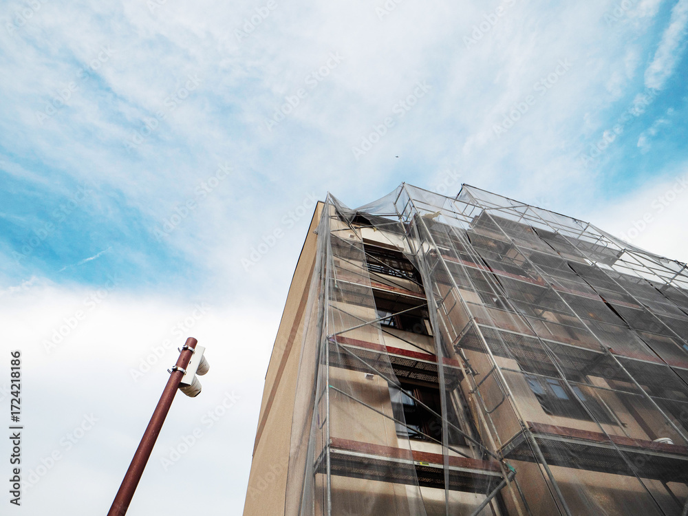 Fototapeta premium Residential apartment building during facade repair or renovation with scaffolding around it in safety mesh. Property upgrade, job increasing value of the home. Construction site.