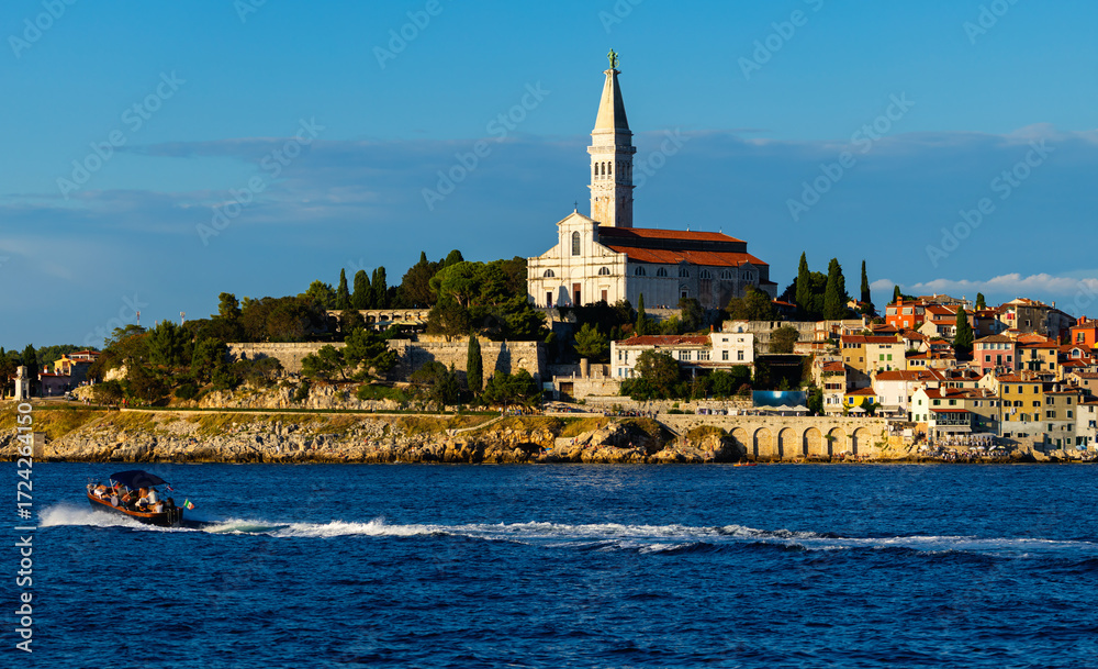 Naklejka premium Rare sailboats and motorboats float past coastal cliffs. Bell tower of Church of St. Euphemia rises above city of Rovinj, citys dominant feature.