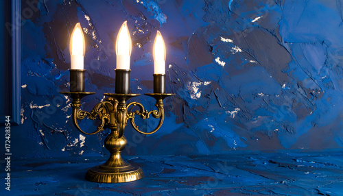 Antique brass three-armed candle holder with illuminated bulbs against a textured deep blue wall.