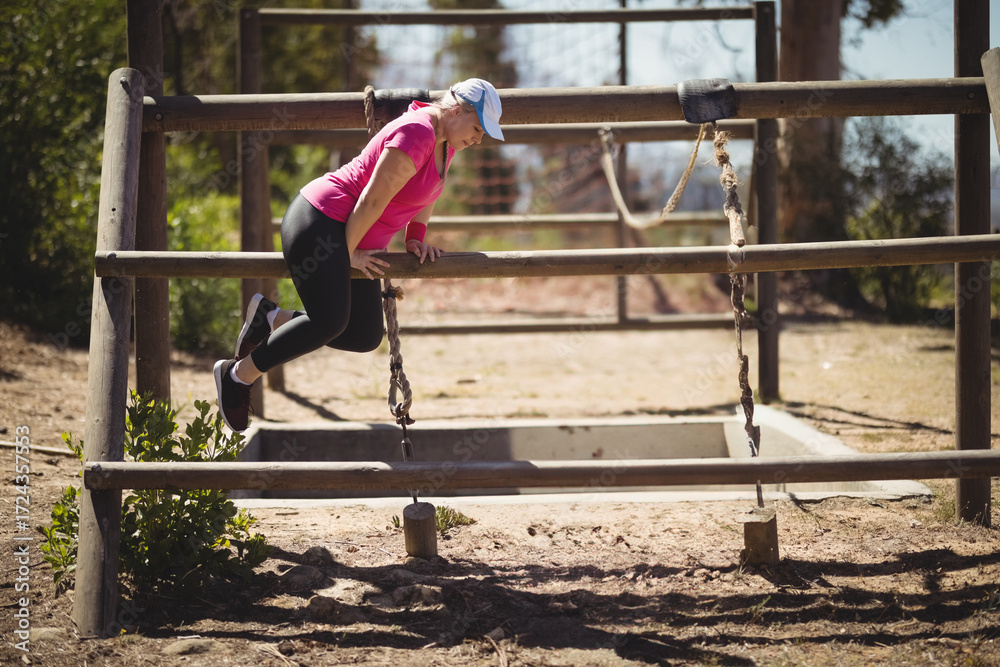 Fototapeta premium Athlete vaulting over wooden three-beam obstacle in sunlit forest clearing with hanging chains