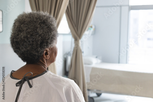 Senior African American patient wearing gown sitting in ward facing bed, viewing window, copy space