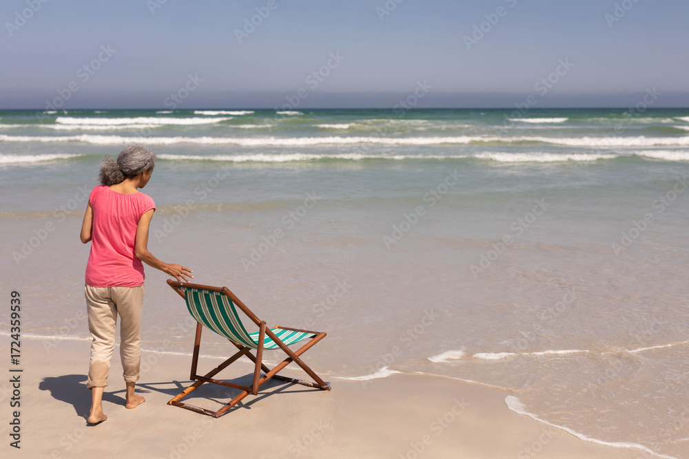Naklejka premium Senior African American woman positioning striped beach chair on wet sand at ocean edge, copy space