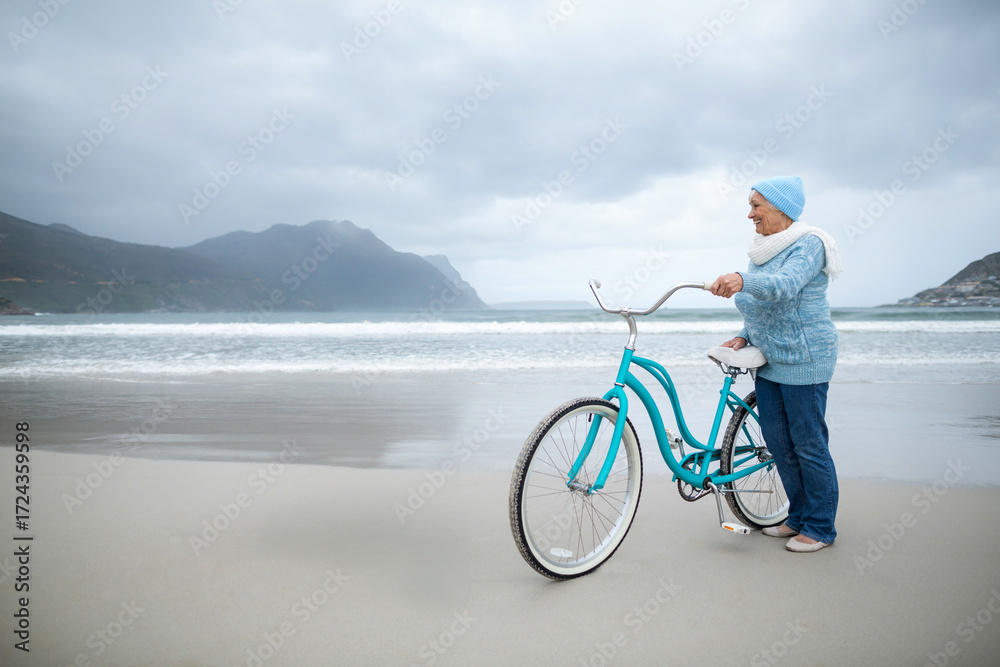 Fototapeta premium Senior woman holding teal bicycle on beach wearing beanie scarf slip-ons gazing at sea, copy space