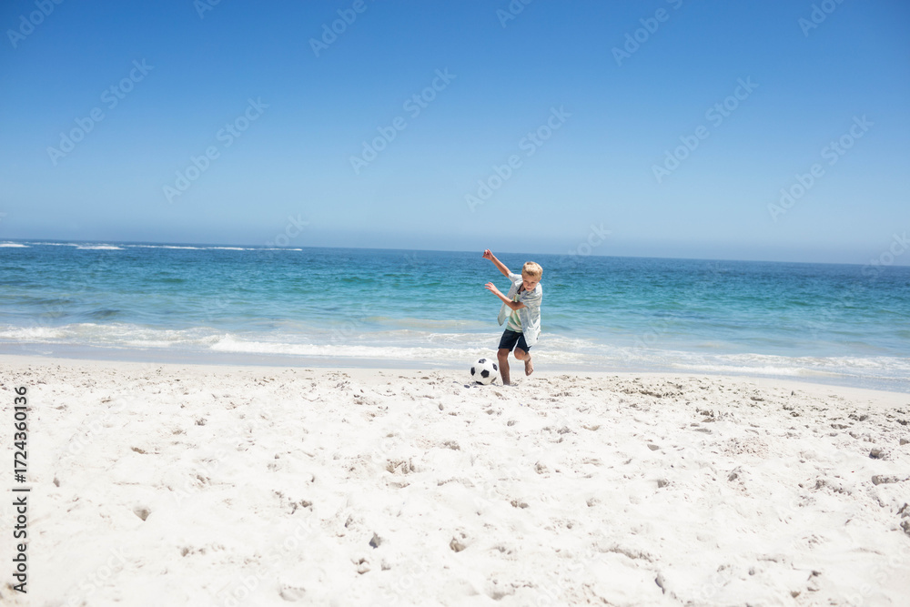 Obraz premium Boy kicking soccer ball at shoreline on white sand near turquoise ocean under clear sky