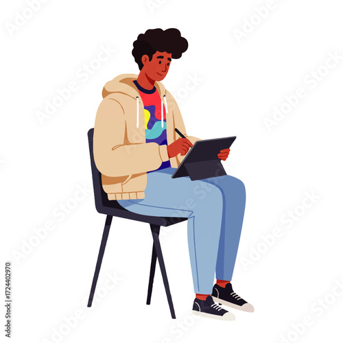 A young man with curly hair sits on a chair, drawing or writing on a tablet with a stylus, wearing a hoodie and jeans.