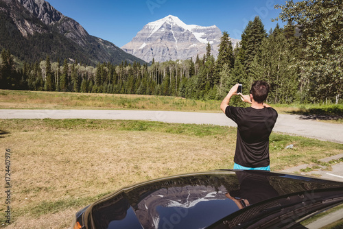 Adult male capturing snow-capped mountain on smartphone by parked car at grassy pullout, copy space