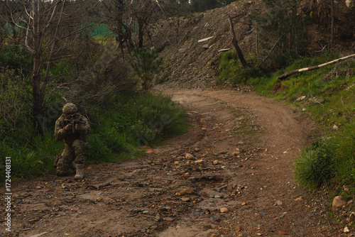 Male soldier kneeling on muddy forest trail along hillside, holding assault rifle, copy space