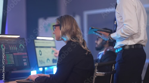 In a modern data analysis center, a female analyst reviews performance dashboards across multiple screens while a manager oversees, collaborating to derive insights from business data.