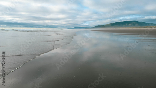 View northward along a beach towards a distant ridge covered in a dark pine forest, with the cloudy and  mysterious sky reflected on the wet sand.
