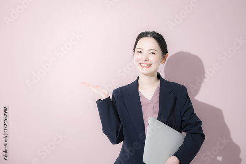 パソコン持ってスーツを着た立つ若いかわいい女性のビジネス上半身　かわいいピンク背景の営業などリクルートのイメージ	紹介や案内、こちら