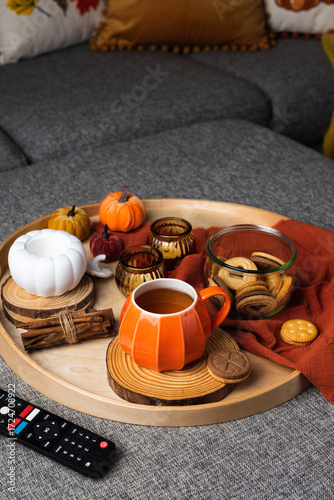 A cozy autumn scene with a tray on a sofa featuring a pumpkin-shaped mug of coffee, cookies in a glass jar, and decorative candles. Warm pillows and a blanket create a relaxed seasonal atmosphere.