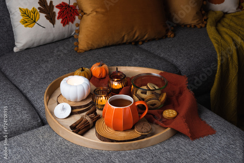 A cozy autumn scene with a tray on a sofa featuring a pumpkin-shaped mug of coffee, cookies in a glass jar, and decorative candles. Warm pillows and a blanket create a relaxed seasonal atmosphere.