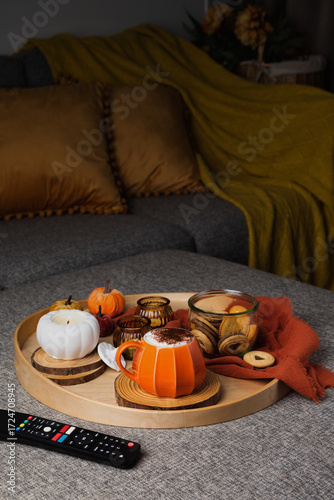A cozy autumn scene with a tray on a sofa featuring a pumpkin-shaped mug of coffee, cookies in a glass jar, and decorative candles. Warm pillows and a blanket create a relaxed seasonal atmosphere.
