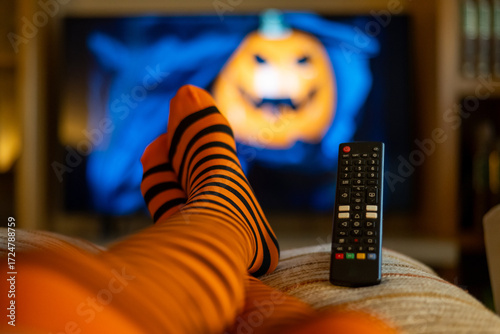 Person relaxing on a couch at home, wearing cozy orange and black striped socks while watching a halloween movie with a blurred jack-o'-lantern visible on the television screen