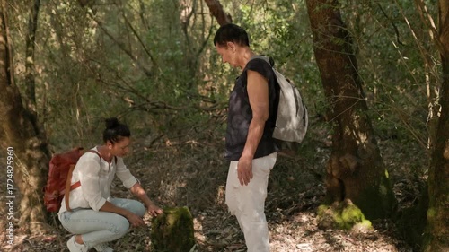 Young woman in white blouse and jeans crouching near mossy stump in forest, while senior woman with gray backpack stands nearby, symbolizing curiosity, connection, and environmental awareness.