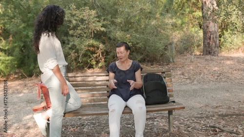 Senior woman in navy blouse with white pants gesturing while sitting on wooden bench, younger woman in white blouse and jeans listening attentively, representing social connection and leisure outdoors
