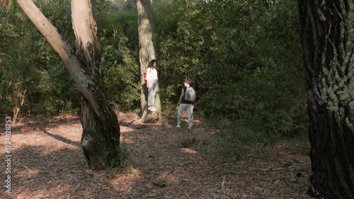 Two women in casual outfits exploring sunny woodland, one carrying red backpack and other with gray backpack, symbolizing outdoor adventure, leisure, and friendship.