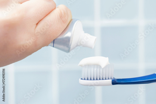 Female hand squeezing toothpaste onto toothbrush against blue tile background.