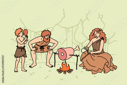 Family of primitive people stands around fire in forest, roasting meat for dinner or sewing up clothes. Group of primitive people of different ages living in stone age dressed in rags