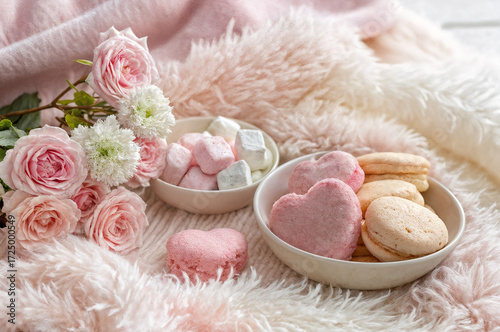 cozy scene with a pink blanket, a bowl of heart-shaped pink marshmallows and cookies, a bowl of white marshmallows, and a bouquet of pink and white roses.