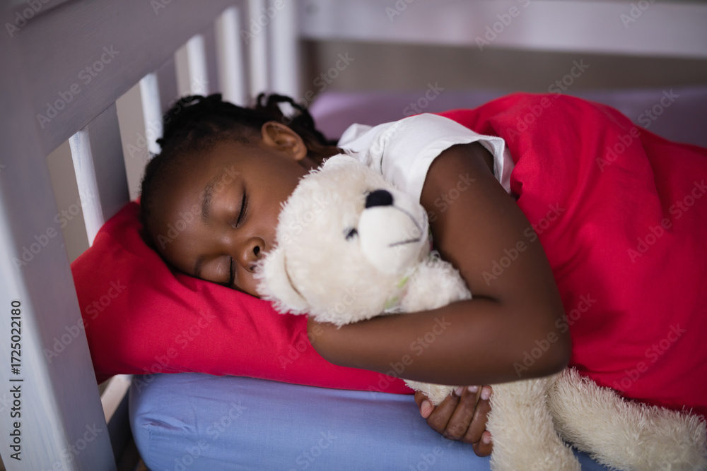 Obraz premium African American child sleeping in bedroom on bed frame hugging white teddy bear under red blanket