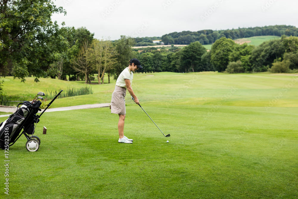 Obraz premium Senior female golfer standing on fairway addressing golf ball with club and push cart, copy space