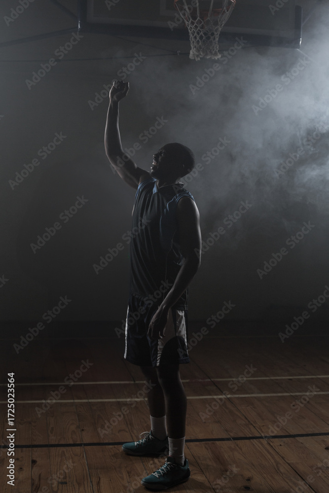 Fototapeta premium Lone athlete standing under basketball hoop on misty wooden court floor, casting subtle shadows
