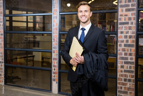 Lawyer standing before glass office wall, holding legal gown and manila folder by brick pillars