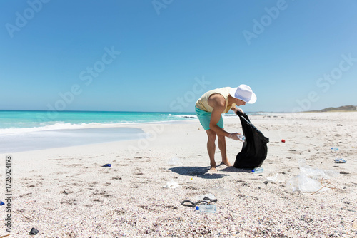 Male volunteer wearing sun hat picking up plastic bottle on beach into trash bag, copy space