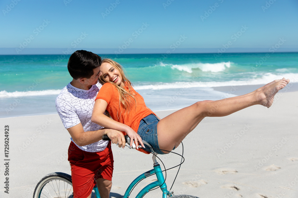 Fototapeta premium Couple balancing teal cruiser bicycle on white sand beach next to turquoise ocean waves