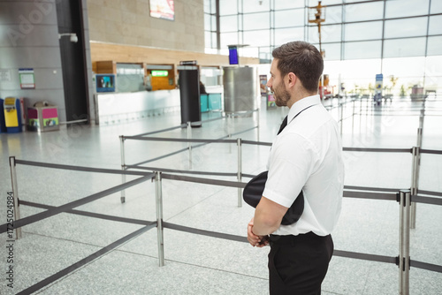 Male pilot wearing uniform standing near queue barriers at airport terminal holding cap, copy space