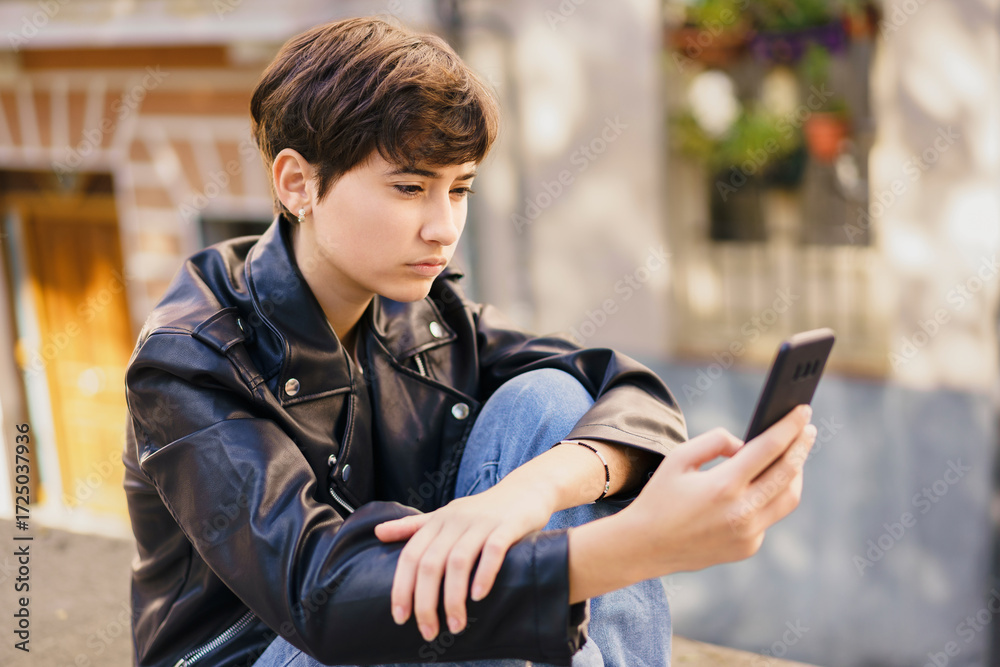Obraz premium A young teenager with a smartphone is seated in an urban setting, surrounded by a lively cityscape
