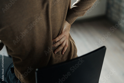 Fototapeta Middle-aged man sitting on a chair holding his lower back in pain, suffering fro