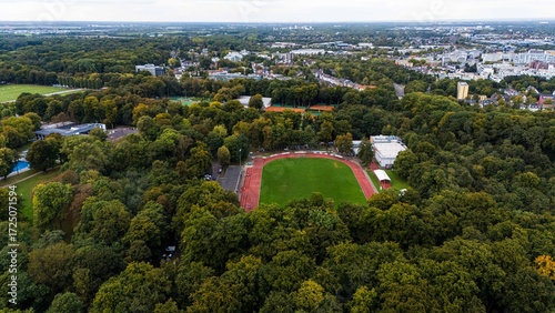 Aerial view of an oval athletics track and football field nestled in a forested urban park. Red running lanes, green turf, and surrounding trees. Drone landscape, daylight, no people.