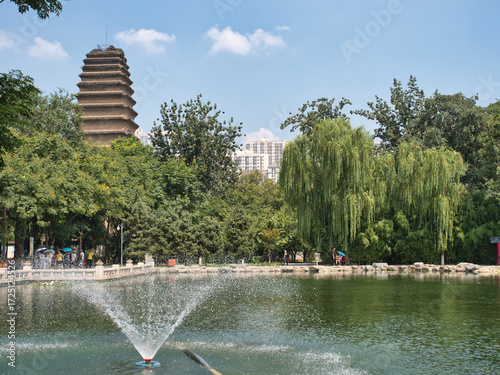 The Giant Wild Goose Pagoda stands majestically behind a tranquil pond and lush trees in a park, with modern city buildings visible in the background
