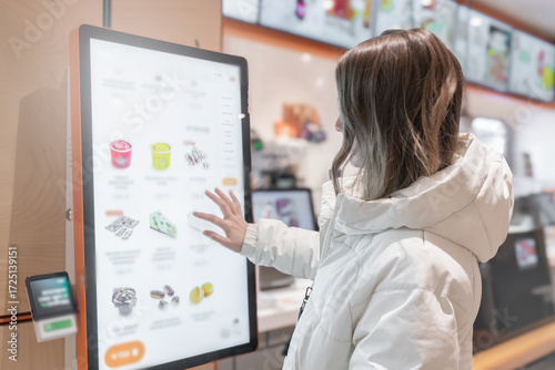 A 30 - 35-year-old woman in a white winter jacket orders cakes and food through a touchscreen screen in a fast food restaurant or pizzeria coffee shop. Person Using Interactive Kiosk for Ordering in a