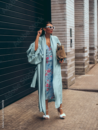 Bild auf Leinwand Fashionable woman in light blue kimono dress and floral print midi dress, holding handbag, posing outdoors with sunglasses