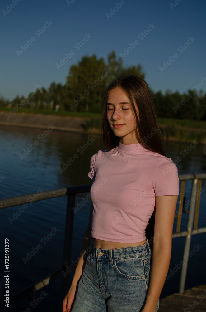 Fototapeta premium A young female model poses outdoors near a lake, enjoying the warm sunlight. She wears a fitted pink top and light blue jeans, with long brown hair flowing down. 