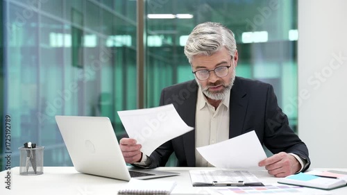 Confused puzzled mature businessman having difficulty with paperwork sitting at workplace in business office. Frustrated financier unhappy with bad financial results holding and reviewing documents