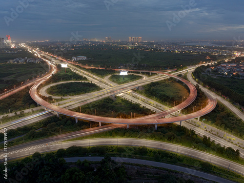 An aerial, long-exposure shot of a cloverleaf interchange in Hanoi at night, with the light trails of moving cars illuminating the complex network of roads surrounded by a cityscape.